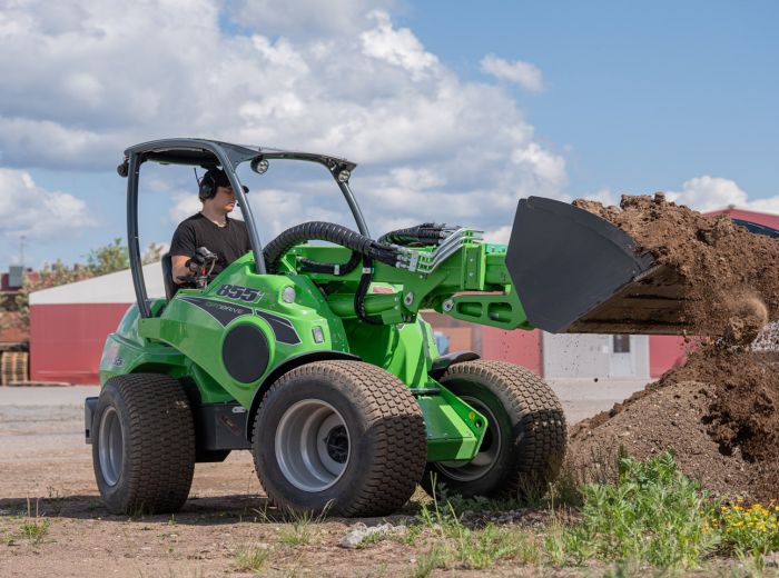 Ein Mann steuert einen grünen Traktor über einen Schotterweg, mit einem Avant Bagger in der Nähe