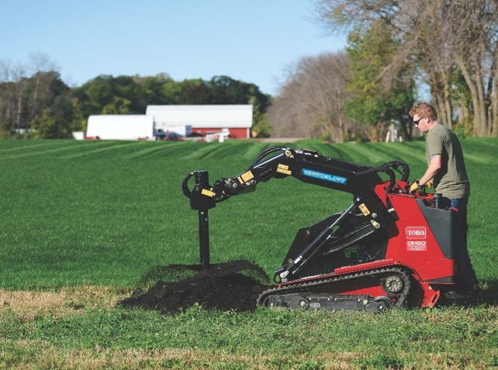 Ein Mann bedient einen kleinen roten Dingo TX1000 Bagger, um im Gras zu graben