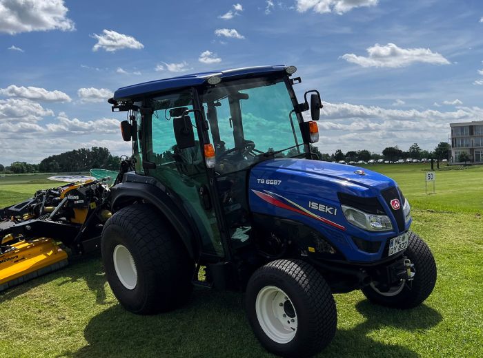 Auf dem Gras geparkter blauer Iseki TG6687 Muething Traktor, neben einem gelben Mäher