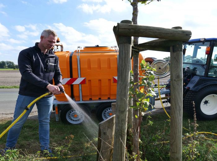 Ein Mann bewässert einen Baum mit einem Schlauch, während er die Bewässerungstechnik von Jansing & Hidding anwendet.