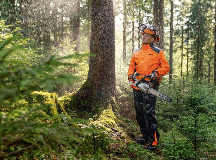 Ein Mann in einer orangefarbenen Jacke steht im Wald neben einer Stihl Motorsäge