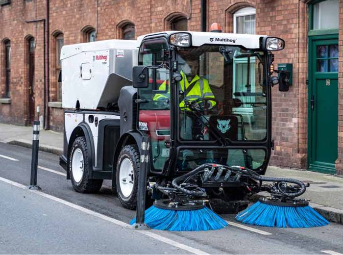 Ein Mann reinigt die Straße mit einem Besen, unterstützt von einer Multihog CV Kehrmaschine im Hintergrund