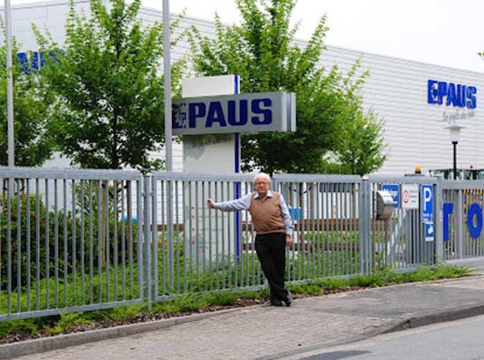 Ein Mann steht vor einem Zaun mit einem Schild, das den Eingang zum Paus Firmengelände anzeigt.