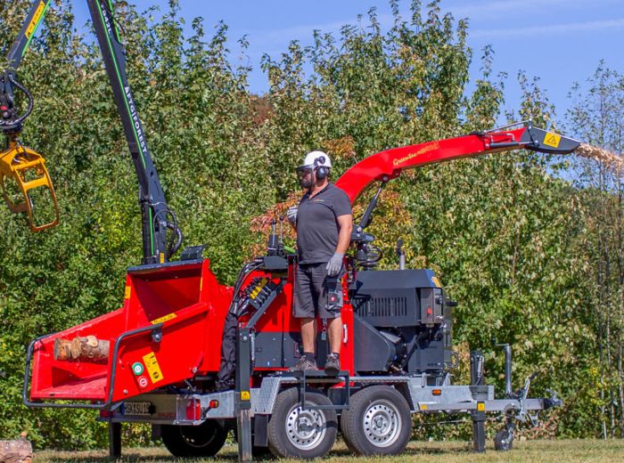Ein Mann steht auf einem roten Anhänger, während ein Baum mit einer Ufkes Maschine gefällt wird.