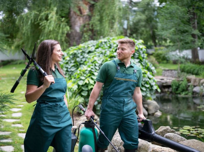 Zwei Personen in grünen Overalls gehen am Ufer eines Teichs entlang, umgeben von Natur und Werkzeugen